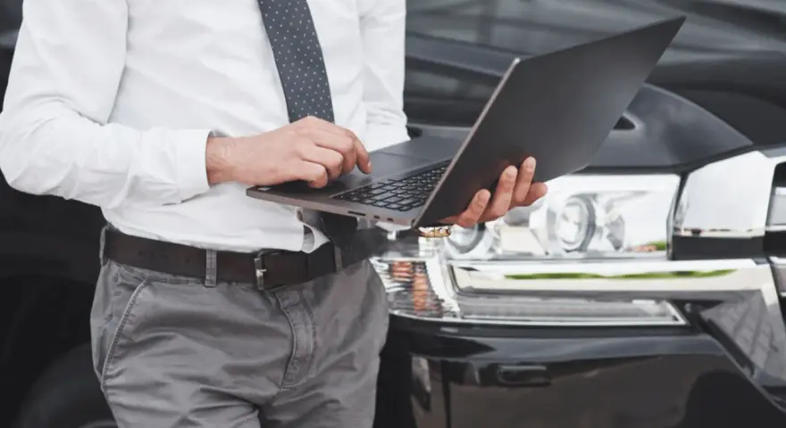 Biznesmen sprawdza na laptopie, ile czasu na przerejestrowanie auta. Elegancki mężczyzna w garniturze przy luksusowym samochodzie.