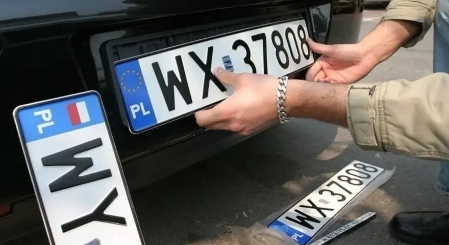Montaż nowych tablic rejestracyjnych. Jedna z nich, z numerem WX 37808, jest właśnie wkładana. Obok leży kolejna, jeszcze w folii.