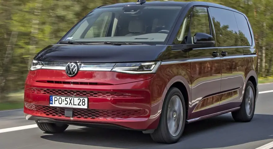 Nowy Volkswagen van w kolorze czerwono-czarnym pędzi po drodze, otoczony zielenią drzew.