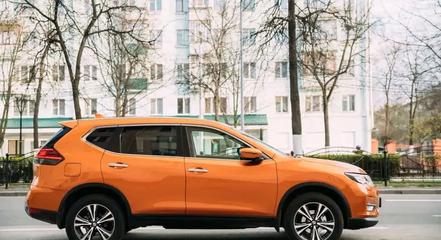 Pomarańczowy SUV do 50 tys. zaparkowany na tle białego budynku i drzew.