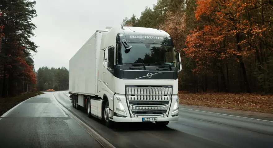 Biała ciężarówka Volvo FH12 z naczepą jedzie po mokrej drodze wśród jesiennego lasu.
