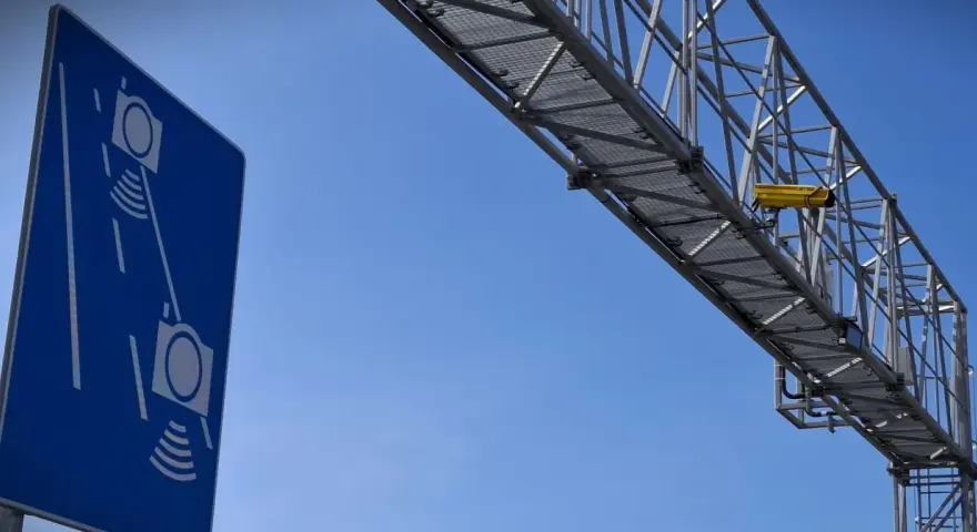 Niebieski znak informuje o odcinkowym pomiarze prędkości S7. Nad drogą widać konstrukcję z żółtą kamerą.