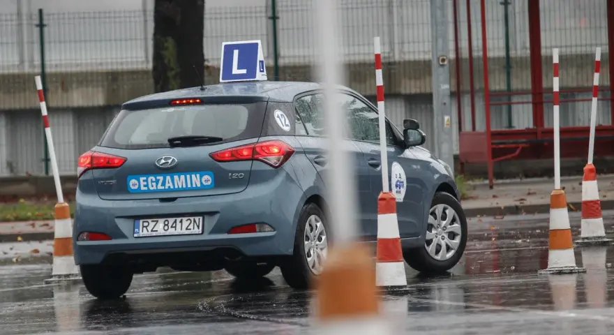 Samochód egzaminacyjny z literą "L" na dachu i napisem "EGZAMIN" na tylnej klapie, otoczony pachołkami na mokrym placu.