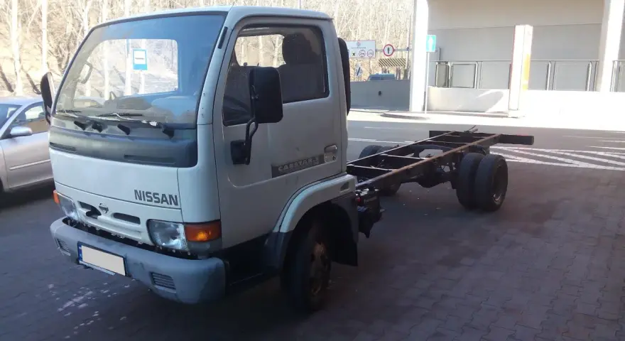 Biały Nissan Cabstar bez zabudowy, z widocznym gołym podwoziem i podwójnymi tylnymi kołami, stoi na kostce brukowej.