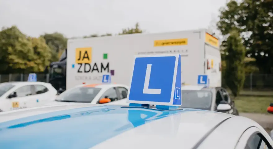 Na dachu samochodu nauki jazdy widoczny znak "L". W tle ciężarówka z napisem "JA ZDAM". Ile jest pytań na kat B?
