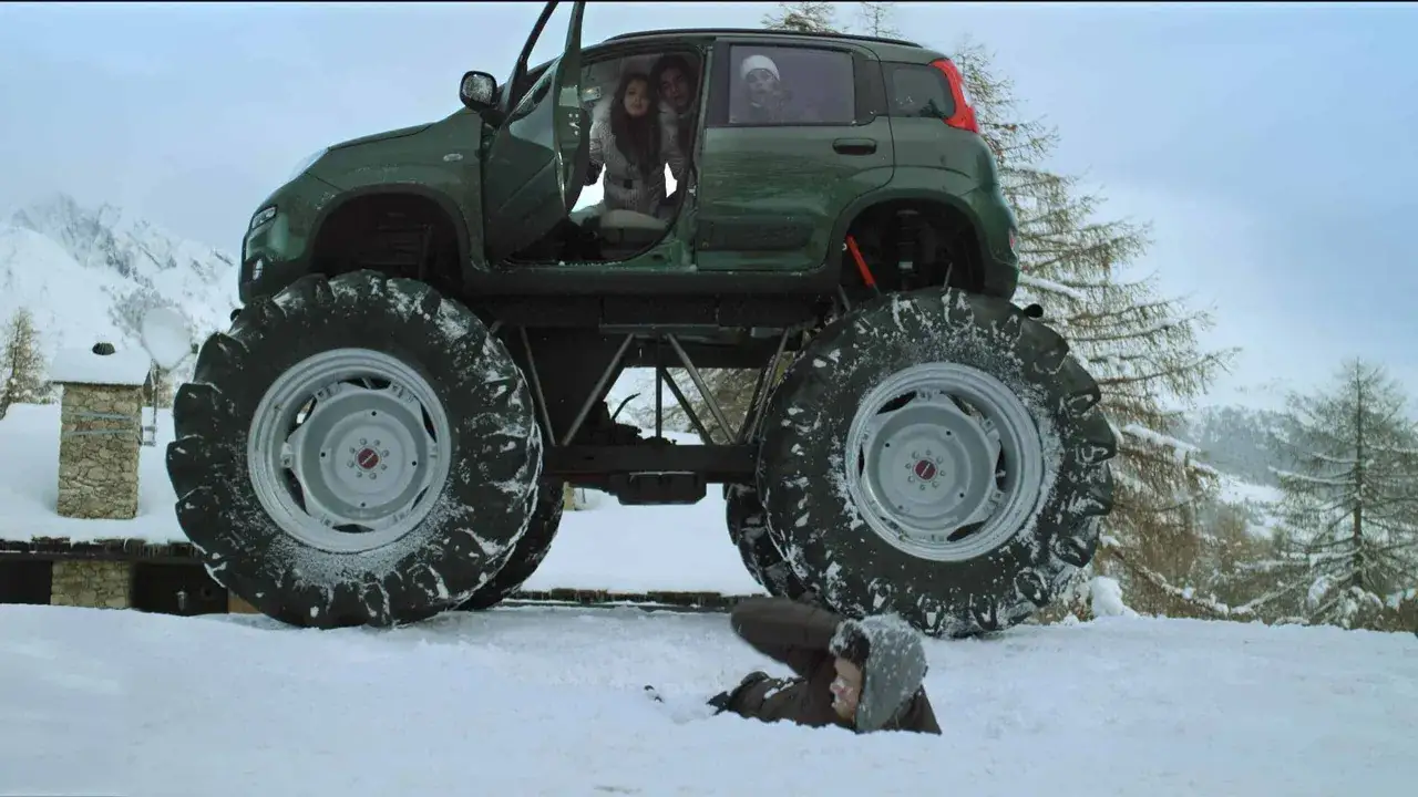 Zielony fiat 4x4 z ogromnymi kołami w śnieżnym krajobrazie. Ludzie w środku, jeden człowiek leży w śniegu.