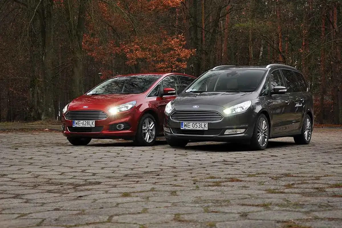 Dwa samochody Ford, czerwony S-Max i szary Galaxy, stoją obok siebie na brukowanej nawierzchni, w tle jesienny las.