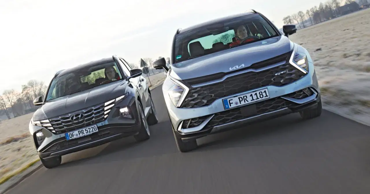 Dwa SUV-y, jeden ciemny Hyundai, drugi jasny suv kia, jadą obok siebie po drodze.