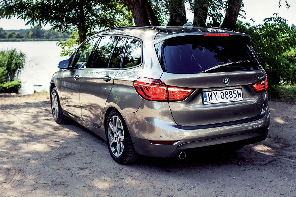 Szary minivan BMW zaparkowany na piaszczystej drodze, z widokiem na jezioro i drzewa w tle.