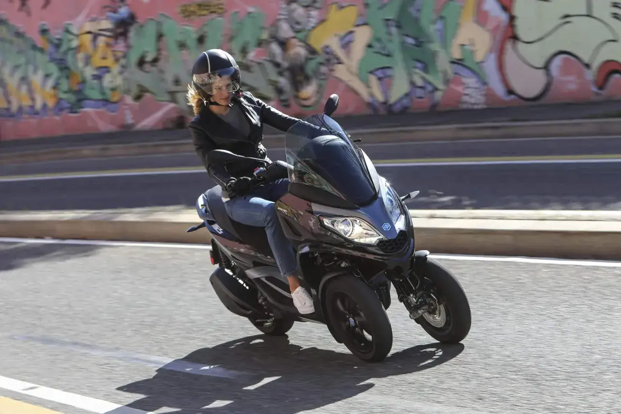 Kobieta w kasku i skórzanej kurtce jedzie na motocyklu trójkołowym na kat B. Tło to kolorowy mural.