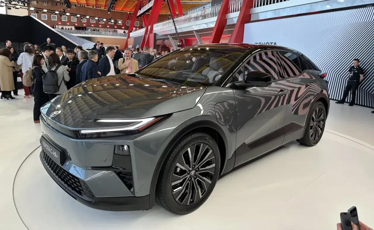 Nowy, szary Toyota C-HR+ na wystawie, otoczony przez ludzi. Elegancki design i nowoczesne linie podkreślają jego sportowy charakter.