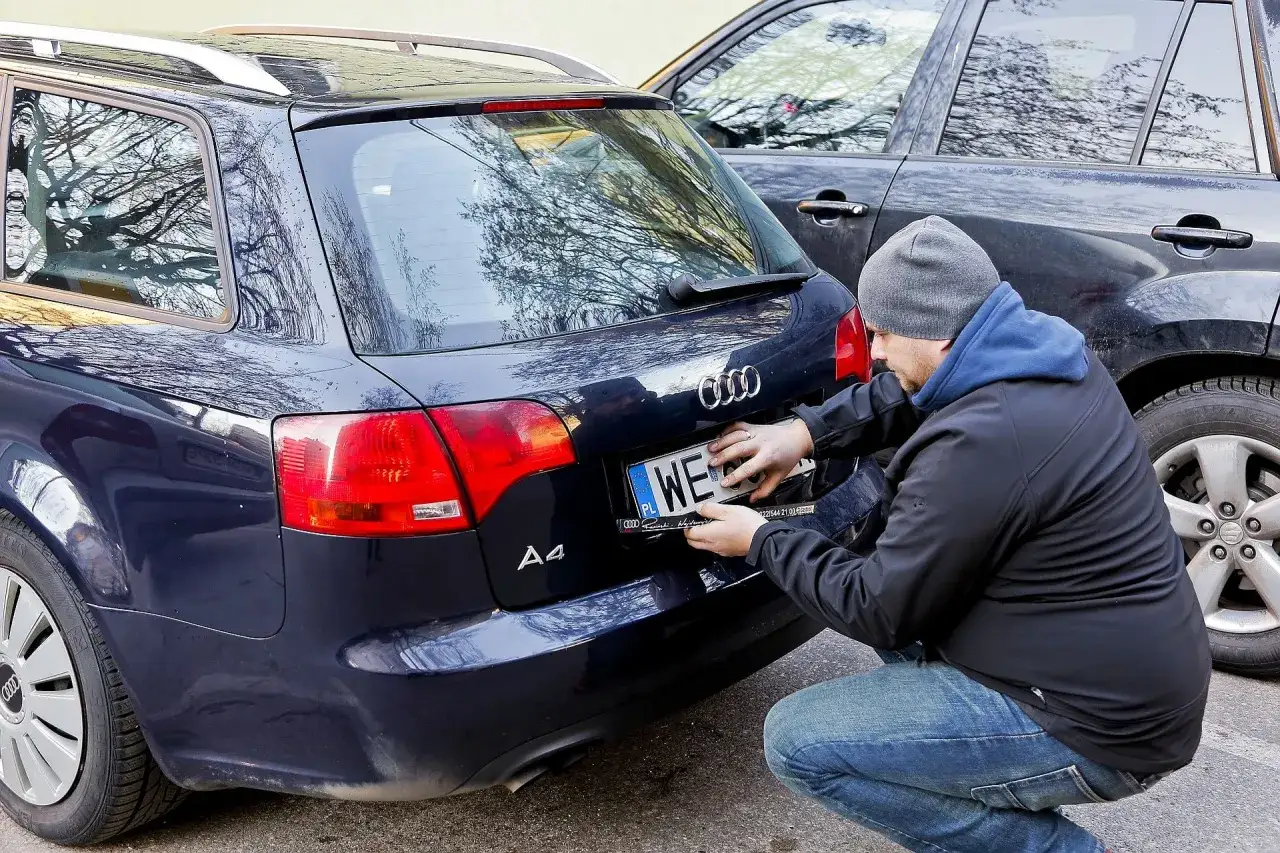 Mężczyzna przykręca zgubioną tablicę rejestracyjną do ciemnego Audi A4.