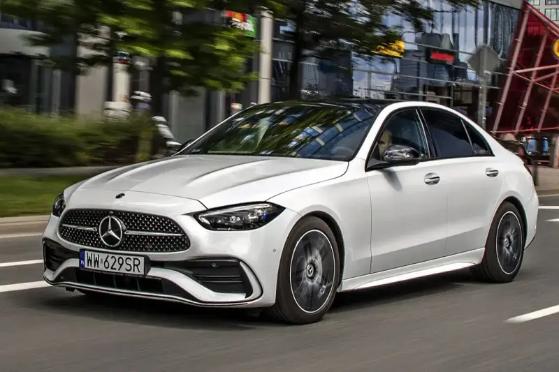 Biały Mercedes C 200 z czarnym dachem sunie po miejskiej drodze, odbijając światła budynków w szybach.