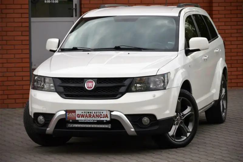 Biały Fiat 4x4 z czarną atrapą chłodnicy i chromowanymi elementami. Auto stoi na kostce brukowej przed budynkiem z cegły.